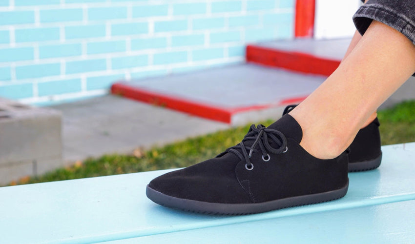 A side view of an Ahinsa Bindu Bare shoe worn by a person sitting on top of a turquoise picnic table