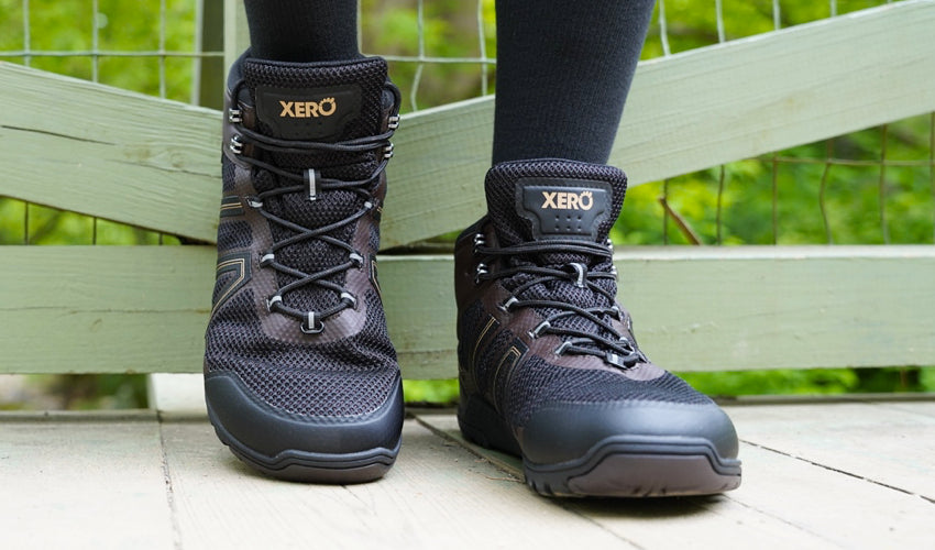 Face-on view of the feet of a person wearing Xero Xcursion Fusion boots with one heel propped up on a fence behind