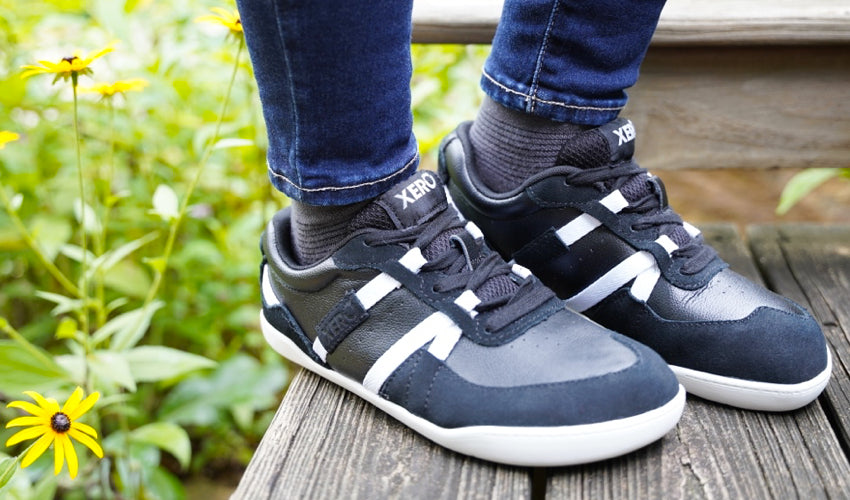 Front-diagonal view of the feet and lower legs of a Xero Kelso shoe-wearer standing on a wooden bench with yellow flowers in the background