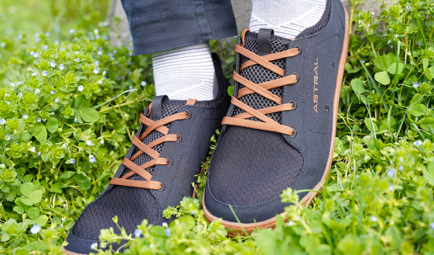 The feet of a person wearing Astral Loyak shoes in Black/Brown in a lush green setting