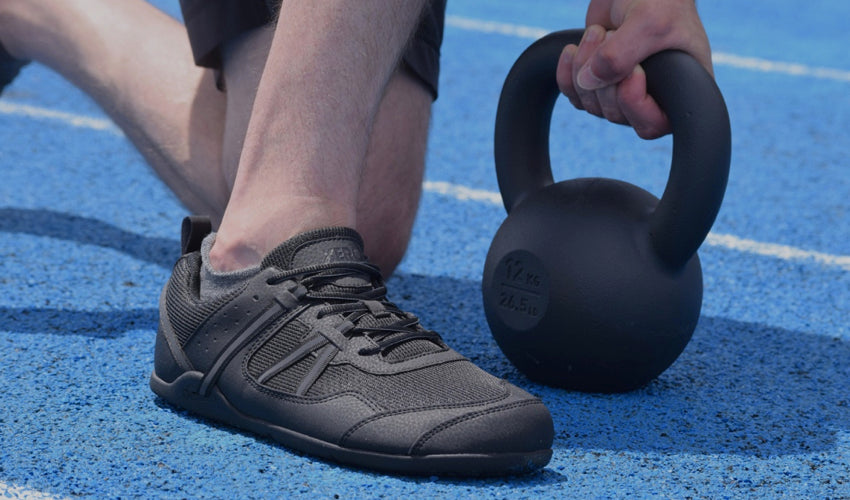 An athlete kneeling on a blue track with a kettlebell and wearing Xero Prio athletic shoes