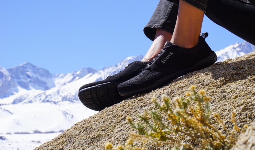 A person wearing Lems Primal 2 Black shoes and sitting on a rocky outcropping with snowy peaks in the background