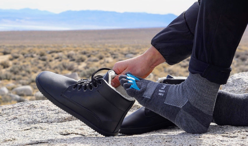 A seated hiker wearing Correct Toes Aqua toe spacers and Injinji Crew Charcoal toe socks donning an Ahinsa Winter Barefoot boot in Black