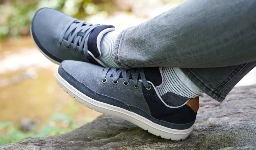 The crossed legs of a person sitting on a rock in nature and wearing a pair of Lems Chillum casual shoes