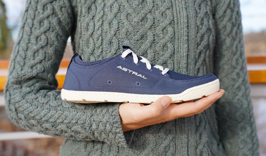 A person, wearing a green-colored wool sweater, holding an Astral Loyak Navy/White shoe in front of her body on an upturned hand