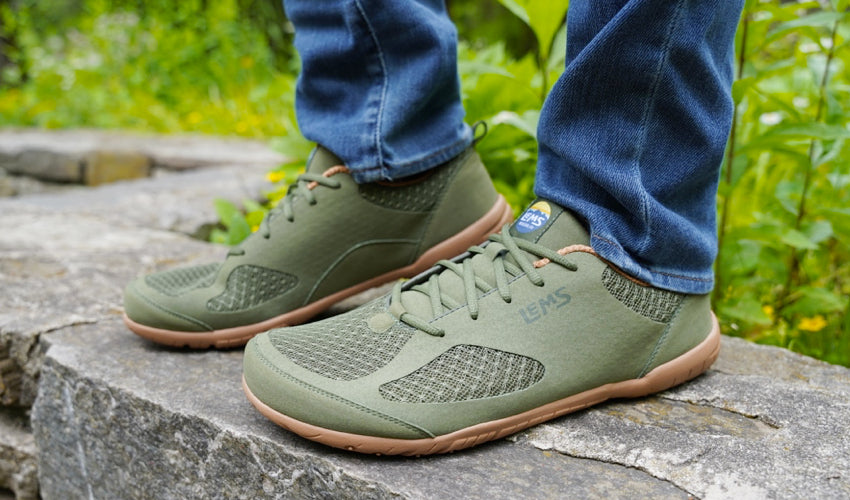 Person wearing Lems Primal 2 Olive shoes and posing on top of a short stone wall