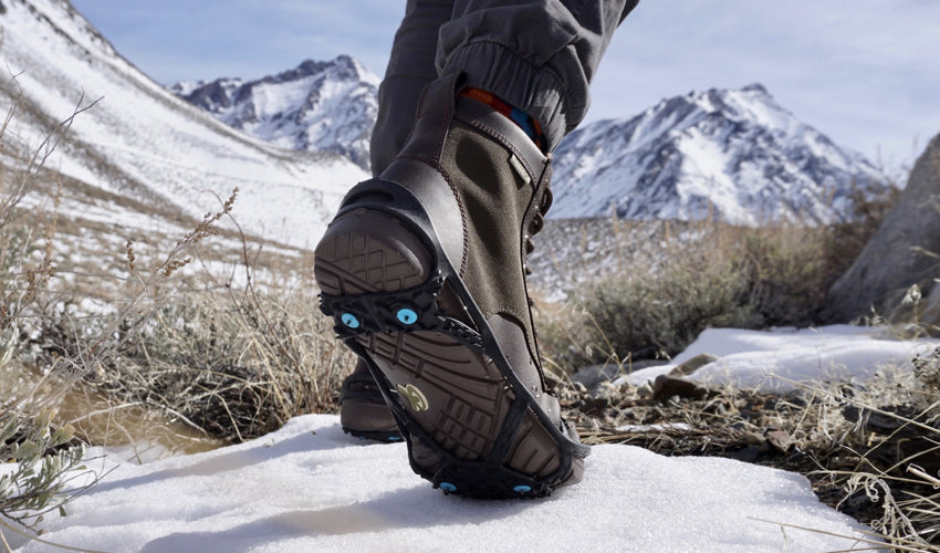 A winter adventurer wearing Lems Boulder Boots and a pair of studded traction aids striding forward into a snowy landscape