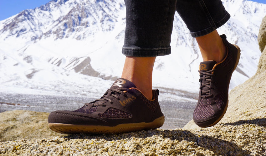Person walking in Lems Primal 2 shoes on a rocky surface with snowy mountains in the background