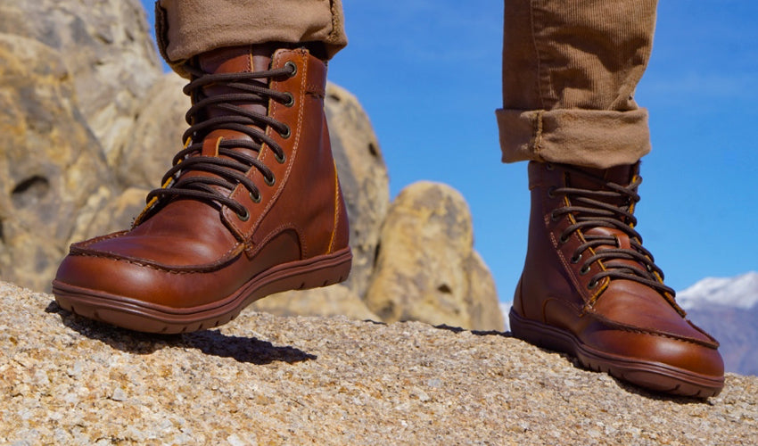 A hiker wearing Lems Boulder Boots in Leather Russet stepping forward with mountains in background