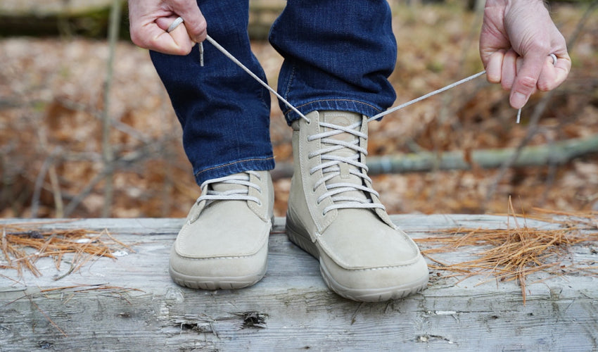 Front view of a person standing on top of a wooden bench and tying the laces of their Lems Telluride Boot