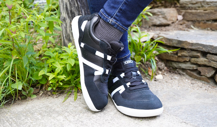 The crossed lower legs of a person wearing Xero Kelso shoes and leaning back against a wooden post with greenery in the background