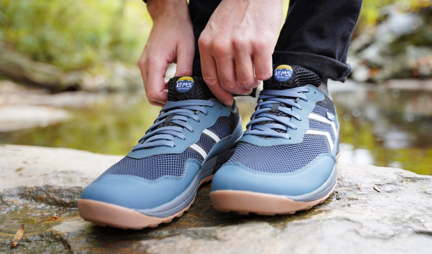 Person standing next to a creek and lacing up a Lems Primal Pursuit Orion Blue shoe