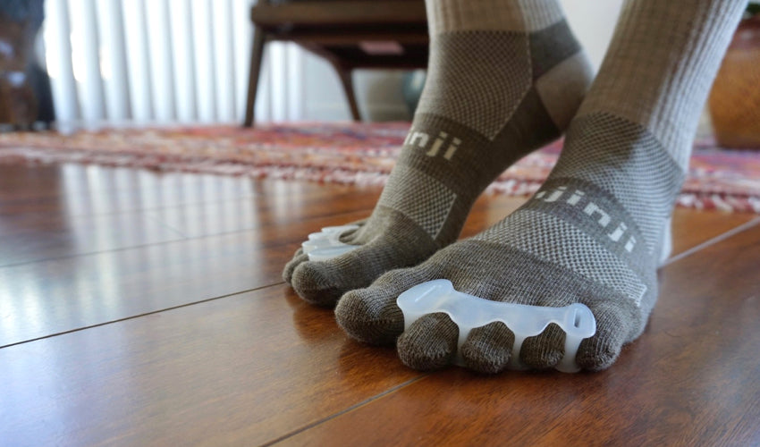 A close-up shot of a person wearing Injinji toe socks and Correct Toes toe spacers and standing on a hardwood floor
