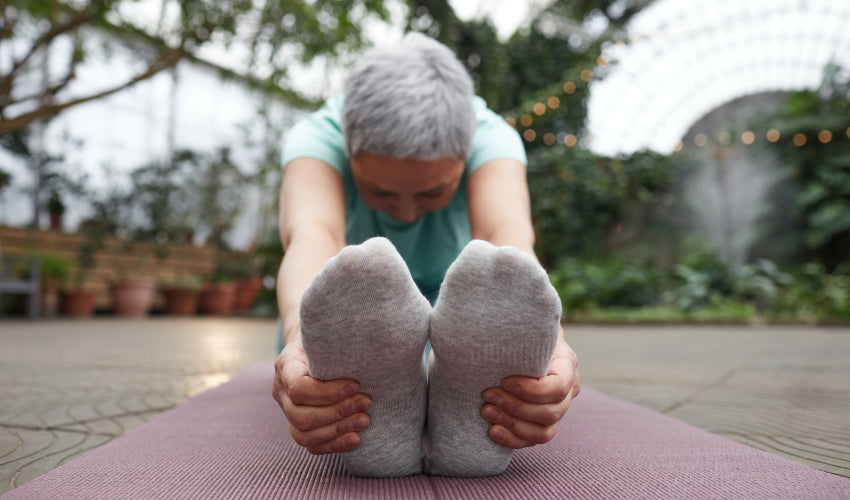 An older individual, seated and with feet in the foreground, leaning forward and stretching on a yoga mat