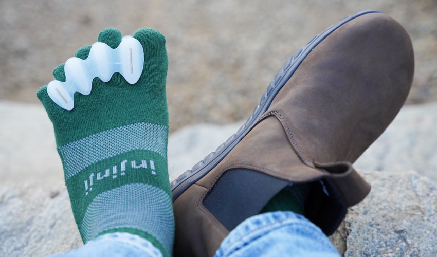 The outstretched legs of a person wearing Correct Toes, Injinji toe socks, and Lems Chelsea Boots in Waterproof Espresso