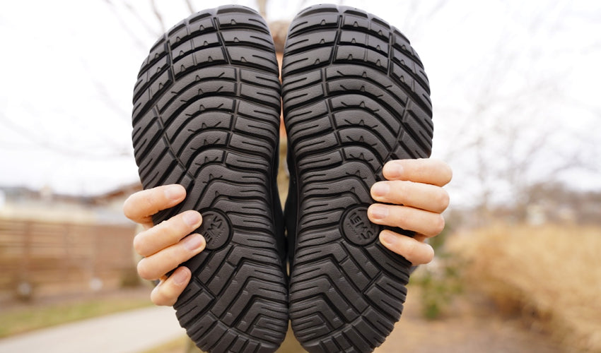 A person with arms outstretched showing the soles of a pair of Lems Chelsea Boot Waterproof Obsidian boots