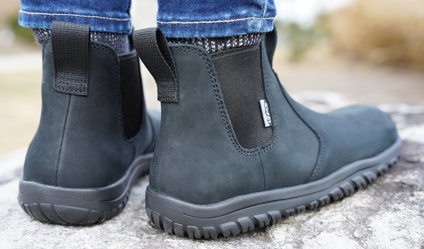 A rear-diagonal shot showing the feet of a standing person wearing Lems Chelsea Boots in Waterproof Obsidian