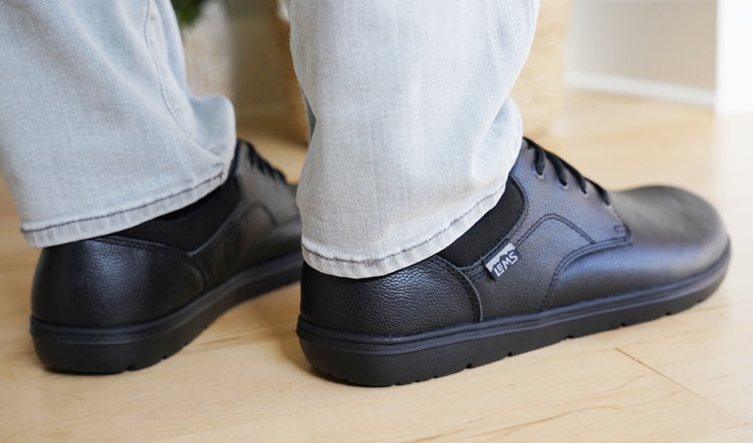 Rear diagonal view of the feet of a person wearing Lems Nine2Five V2 Black shoes on a wood floor