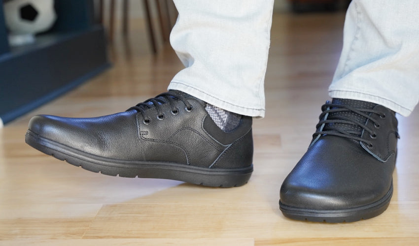 The feet and lower legs of a person wearing Lems Nine2Five V2 Black shoes and standing in a leisurely fashion