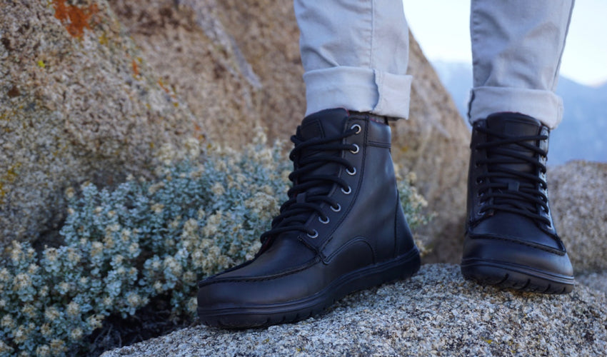 A person posing in a pair of Lems Boulder Boots in Leather Raven with colorful rocks in the background