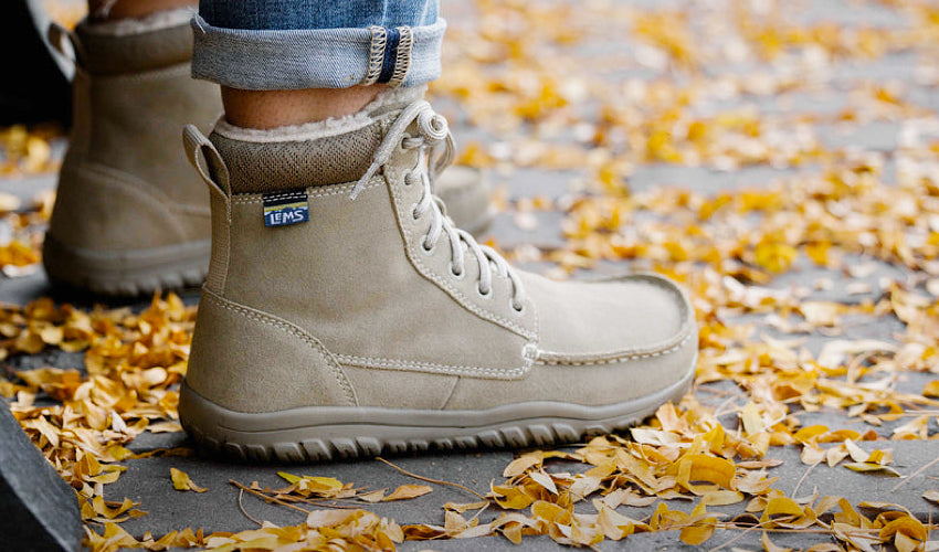 The feet of a seated person wearing Lems Telluride Boots in Limestone with colorful fall leaves surrounding them