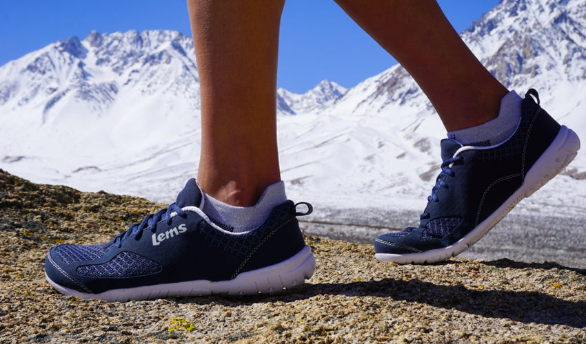 A hiker wearing Lems Primal 2 Eclipse minimalist shoes with snow-capped mountains in the background