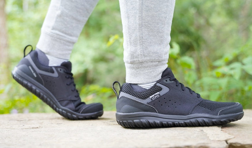 Side view of a person walking atop a stone wall and wearing a pair of Lems Primal Zen Asphalt shoes