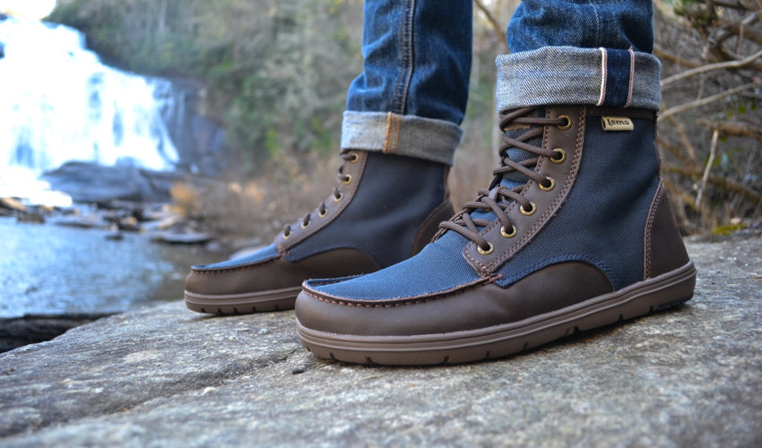 A person standing on a rocky embankment wearing Lems Boulder Boots in Nylon Navy Stout and taking in a waterfall in the background