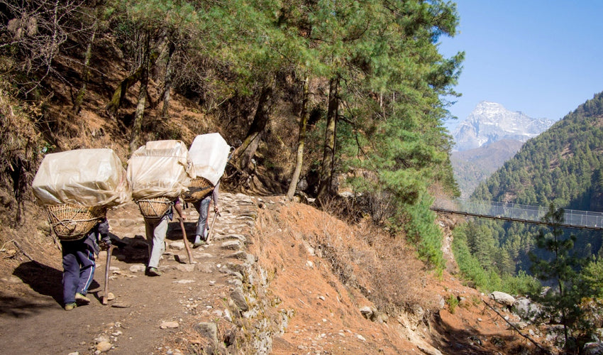 Three Nepali porters walking barefoot in the Himalayas while carrying massive loads on their backs