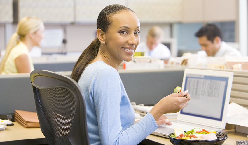 A smiling office worker in a cubicle eating lunch while working on her laptop