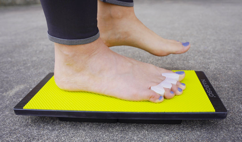 Barefoot person wearing a Correct Toes toe spacer and performing a single-leg stand on a Naboso Kinesis Board