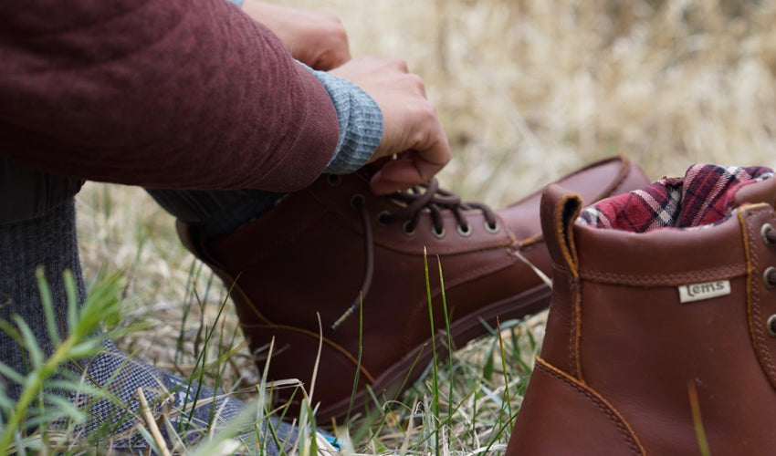 A person seated in a grassy meadow tying the laces of a pair of Lems Boulder Boots in Leather Russet