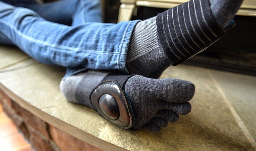 A person seated on a stone hearth with outstretched legs wearing Injinji toe socks and Strutz Pro Black foot pads