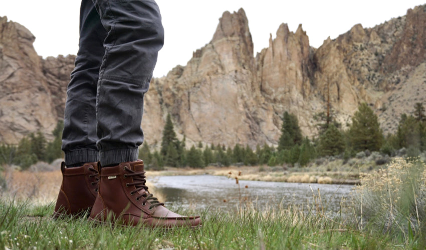 Person wearing the Lems Boulder Boot in Leather Russet and standing in a grassy field with mountains and stream in the background