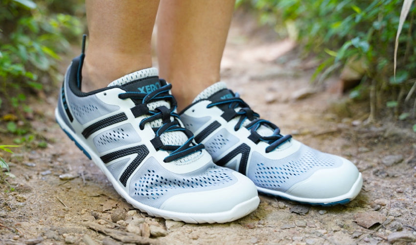 The lower legs of an athlete wearing Xero HFS shoes and posing on a forest trail