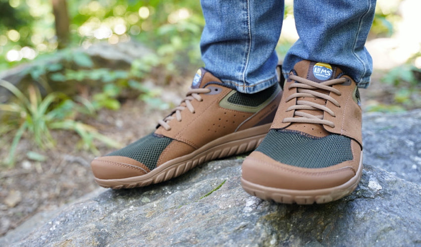 Person wearing a pair of Lems Primal Zen Woodland shoes and standing on a stone in a forest