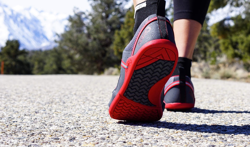 The tread pattern of a pair of Xero Prio athletic shoes worn by a person walking on a chipseal road