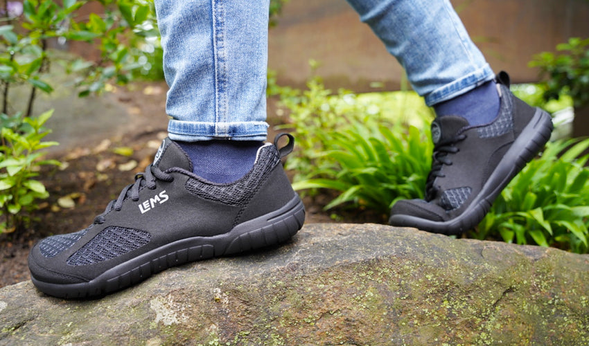 Side view of the feet of a Lems Primal 3 shoe wearer striding over a large stone in a garden setting