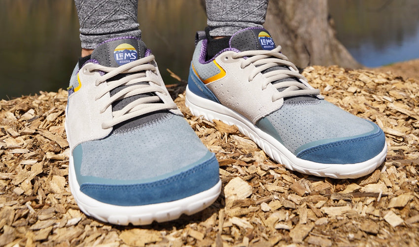 Front view of a person wearing Lems Primal Zen Suede Sun Ray shoes with a river in the background