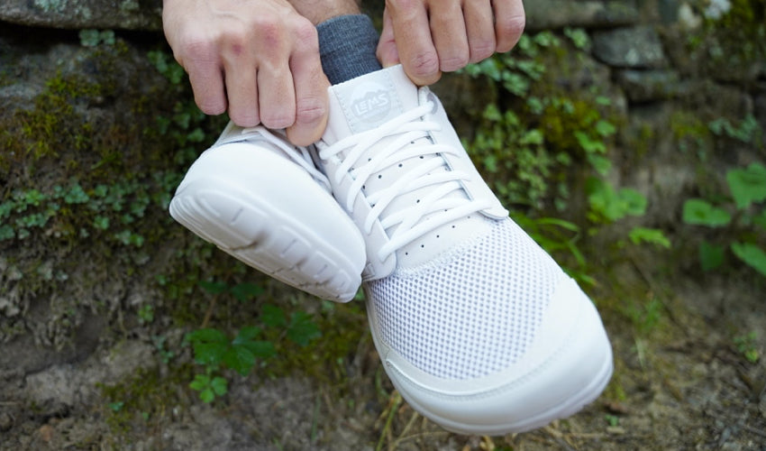 Person sitting on a stone wall and donning a pair of Lems Primal Zen White Sand shoes