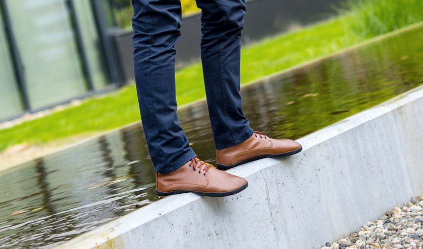 Person wearing Ahinsa Sundara Ankle Barefoot boots and standing on a concrete ledge next to an urban pond