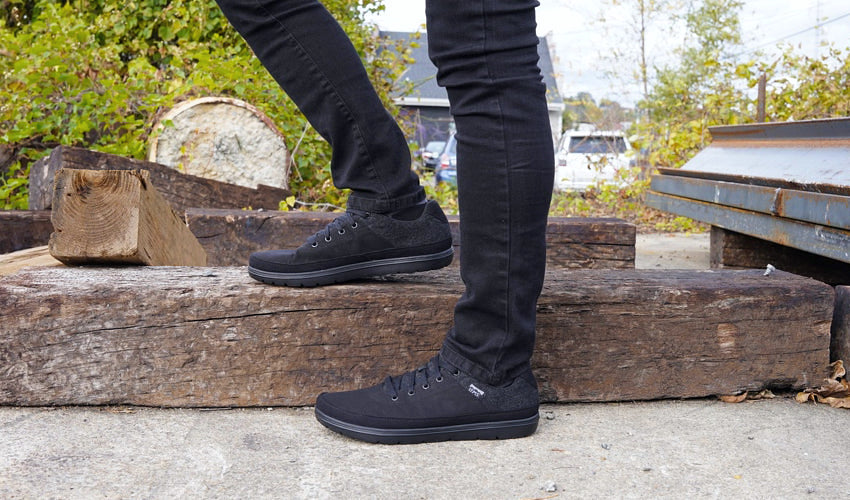 Side view of person in black jeans and Lems Chillum Blackjack shoes posing with an old railroad tie