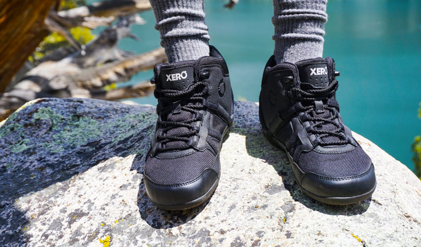 A hiker, standing on a rock and with a green lake in the background, wearing Xero DayLite Hiker EV Black hiking boots