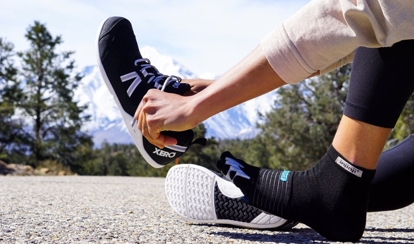 A person, wearing a spate of natural footgear, donning a pair of Xero Prio athletic shoes with snowy mountains in the background