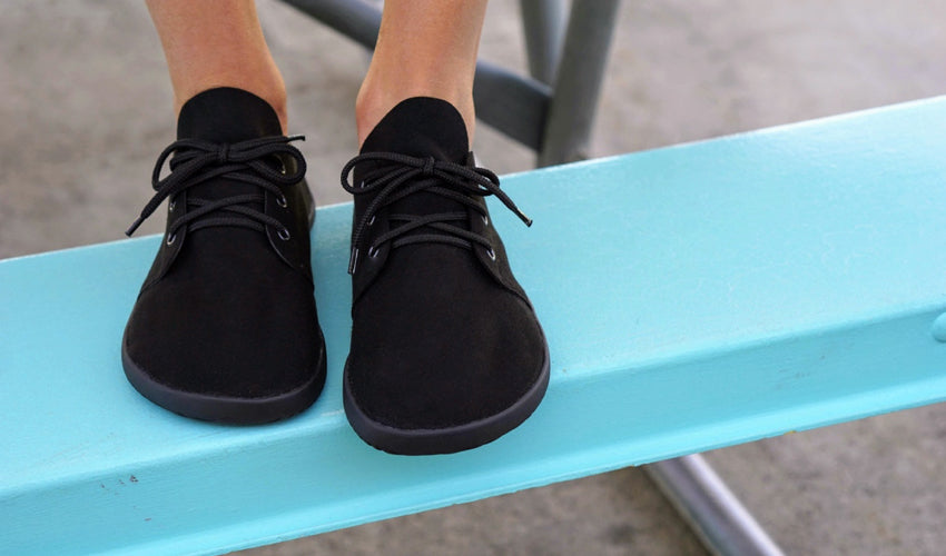 A front-on view of a pair of Black Ahinsa Bindu shoes worn by a person sitting on a turquoise picnic table