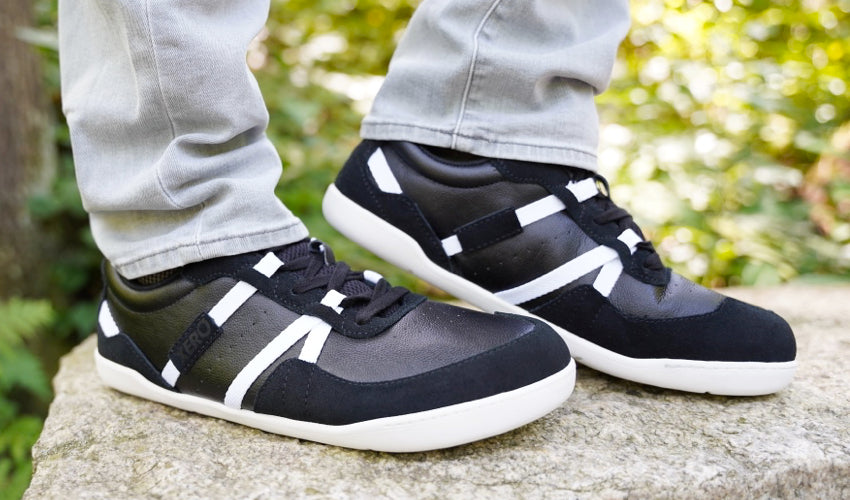 Side view of the feet of a person standing on top of a flat stone bench wearing Xero Kelso shoes in Black/White