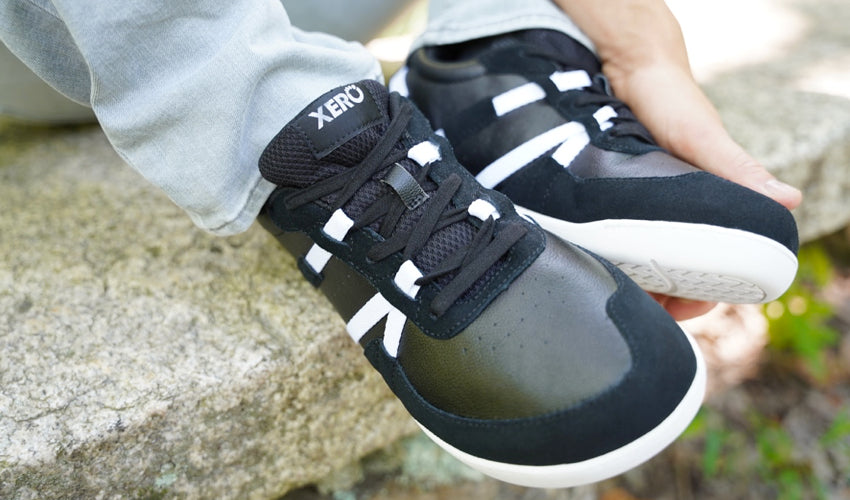 Top-down view of the feet of a seated person wearing Xero Kelso Black/White shoes with shoes positioned side-by-side and supported by the wearer's left hand