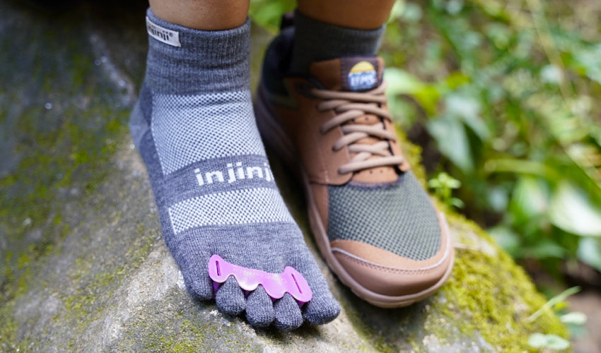 The feet and lower legs of a hiker wearing foot-healthy footwear, toe spacers, and toe socks sitting on a mossy rock in woods