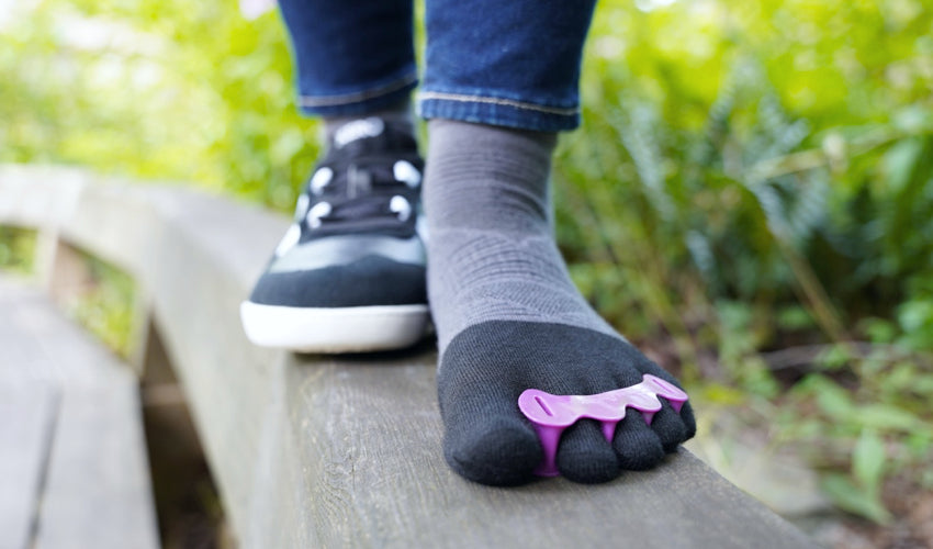 Front view of a person on a wooden bench striding forward, one foot wearing Correct Toes Plum toe spacers and Injinji toe socks, the other foot a Xero Kelso Black/White shoe