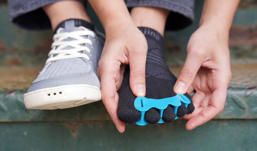 Close-up, head-on view of a person sitting on green metal steps and donning Correct Toes Aqua toe spacers over Injinji toe socks